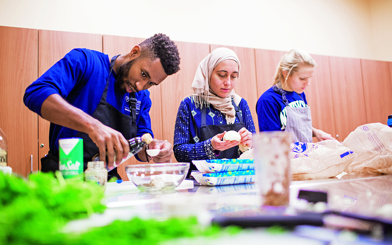 Graduate students dressed in aprons prepare a meal behind a vegetable-laden table.
