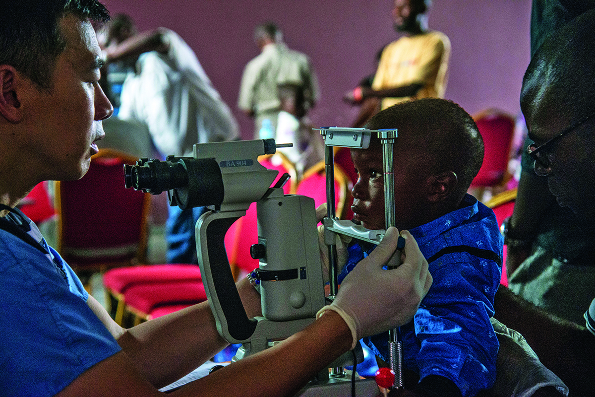 An Emory doctor in blue scrubs uses an optical device to check the eyes of a young boy in the Democratic Republic of Congo.