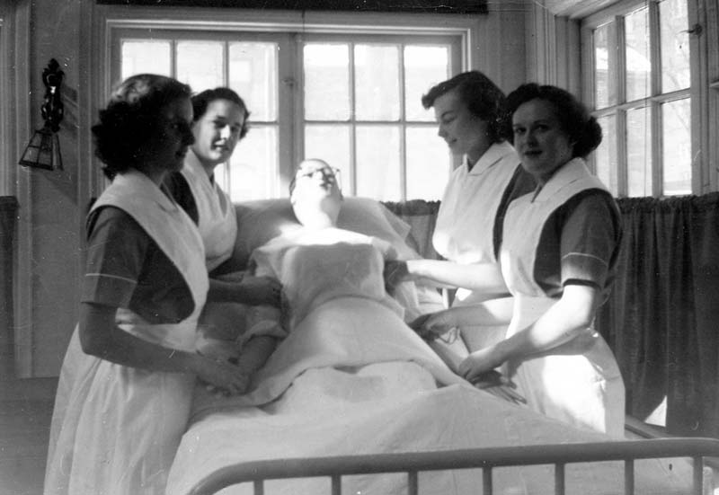 4 nurses in a black and white photo from the early part of the 20th Century stand around a dummy on a bed.