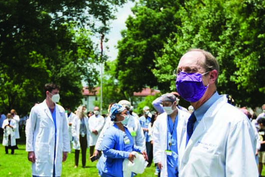 John Lewin and many others, all wearing lab-coats and masks, mill about on a lawn.