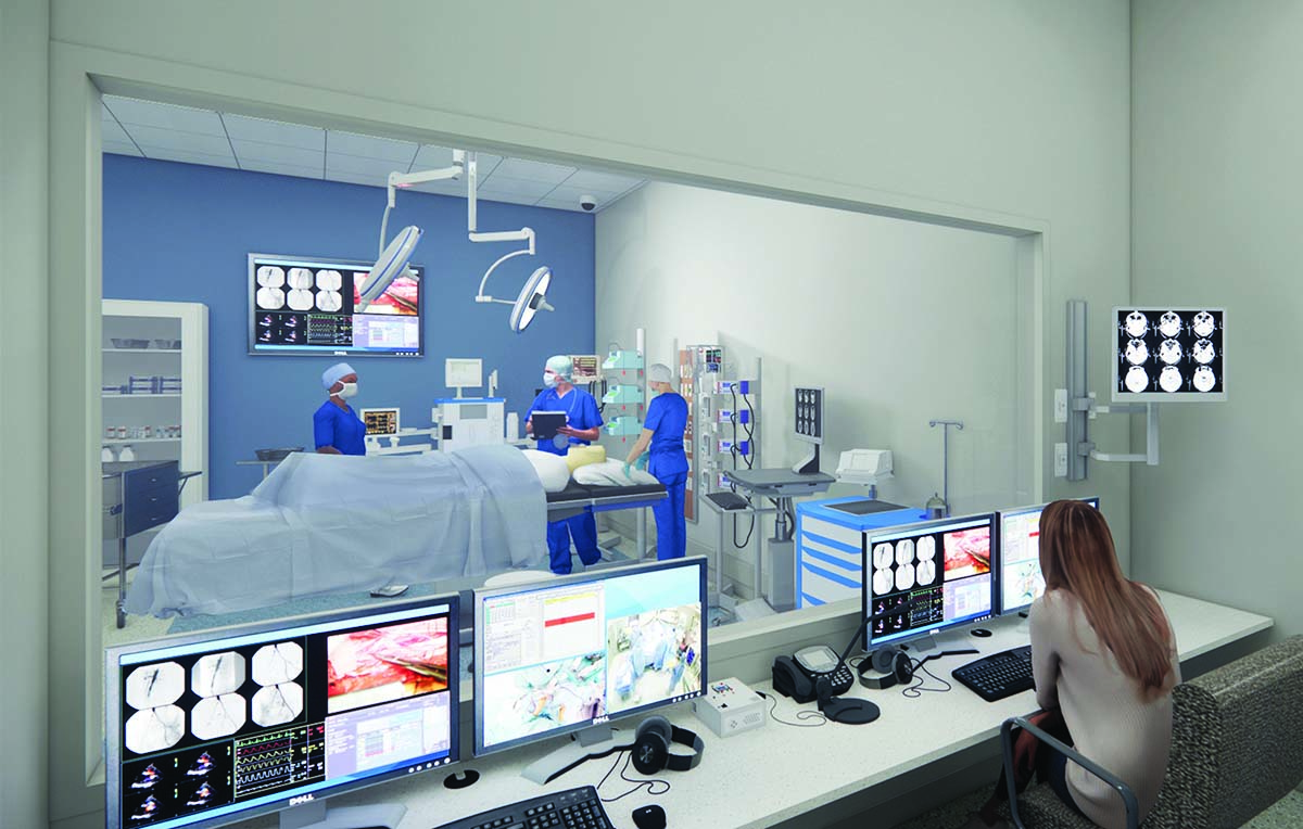 A woman sits with her back to the camera before monitors and a large window. Through the window, she is observing three masked nurses working around a dummy on an exam table.