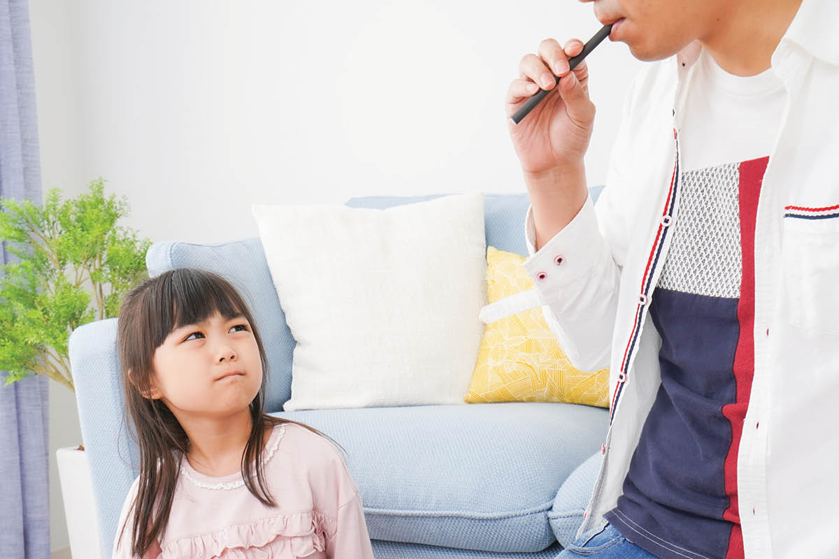 Photo of child sitting at table looking up an adult using an e-cigarette with a disapproving look on her face.